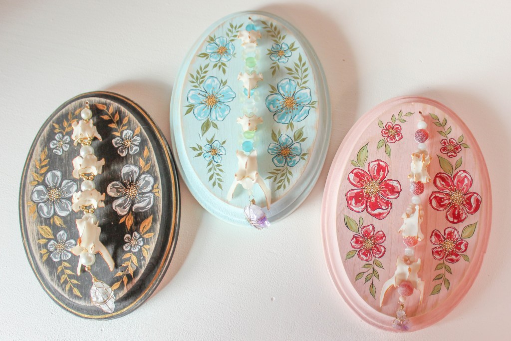 A display of three decorative oval plaques featuring floral designs, each adorned with shells and beads. The left plaque is black with white and gold flowers, the middle one is light blue with blue flowers, and the right one is pink with red flowers.